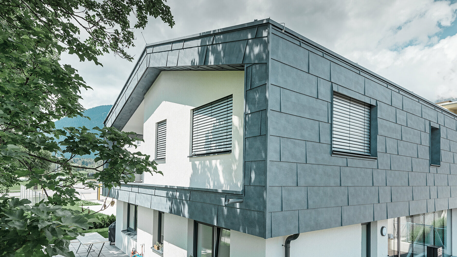 Moderna obiteljska kuća bez strehe; gornja etaža obložena je PREFA fasadnim panelima FX.12 u kameno sivoj, ostatak fasade je bijela žbuka.