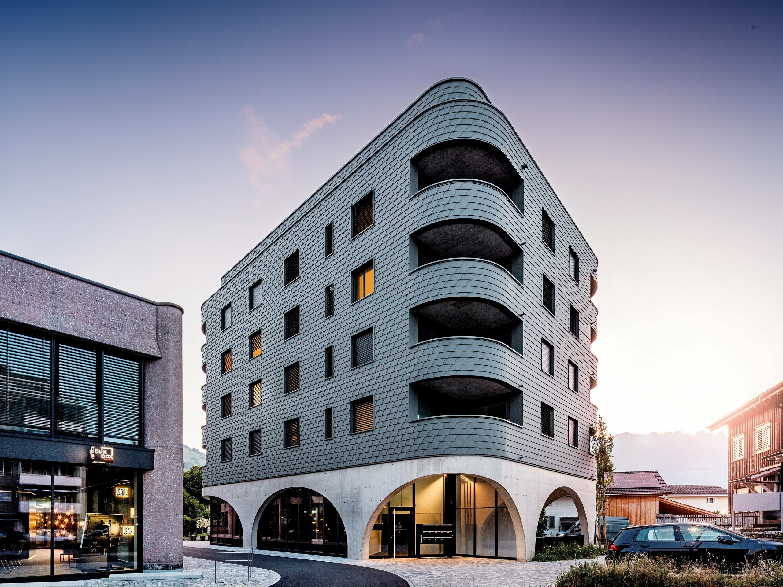 Moderna višefunkcionalna zgrada u Buchsu s upečatljivim zaobljenim, trokutastim oblikom i kontinuiranom fasadom od PREFA zidnih šindra u P.10 svijetlosivoj boji. Ljuskasta aluminijska fasada obavija zgradu poput reptilske kože, naglašavajući tekuće zaobljene linije i duboko urezane balkone. Iznad masivnog postolja s elegantnim betonskim lukovima, lagana, otporna na vremenske uvjete PREFA fasada djeluje posebno izražajno. Precizno postavljene aluminijske šindre stvaraju homogenu, dugotrajnu i gotovo bezdržavnu ovojnicu zgrade s jakim prepoznatljivim karakterom.