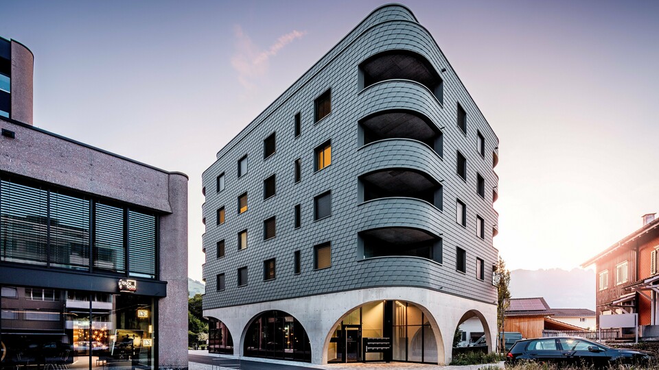 Moderna višefunkcionalna zgrada u Buchsu s upečatljivim zaobljenim, trokutastim oblikom i kontinuiranom fasadom od PREFA zidnih šindra u P.10 svijetlosivoj boji. Ljuskasta aluminijska fasada obavija zgradu poput reptilske kože, naglašavajući tekuće zaobljene linije i duboko urezane balkone. Iznad masivnog postolja s elegantnim betonskim lukovima, lagana, otporna na vremenske uvjete PREFA fasada djeluje posebno izražajno. Precizno postavljene aluminijske šindre stvaraju homogenu, dugotrajnu i gotovo bezdržavnu ovojnicu zgrade s jakim prepoznatljivim karakterom.