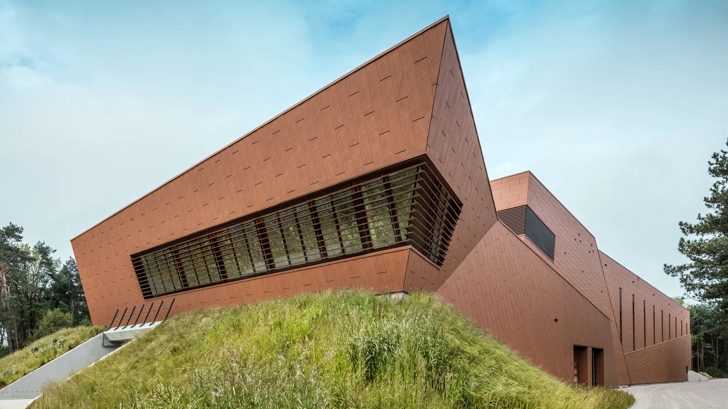 Markantes Trinkwasserpumpwerk von WML in Lottum mit skulptural geneigter Gebäudeform und prägnanter PREFA Siding.X Aluminiumfassade in warmem Rostton. Die dynamisch strukturierte Oberfläche reflektiert Licht und setzt starke architektonische Akzente im Landschaftsraum. Großzügige Lamellenöffnungen und klare Linienführung verbinden Technik und Gestaltung. Die langlebigen, leichten und korrosionsbeständigen PREFA Aluminiumpaneele überzeugen durch nachhaltige Recyclingfähigkeit, hohe und eine wartungsarme Gebäudehülle – ein moderner Eyecatcher für zukunftsorientierten Infrastrukturbau.