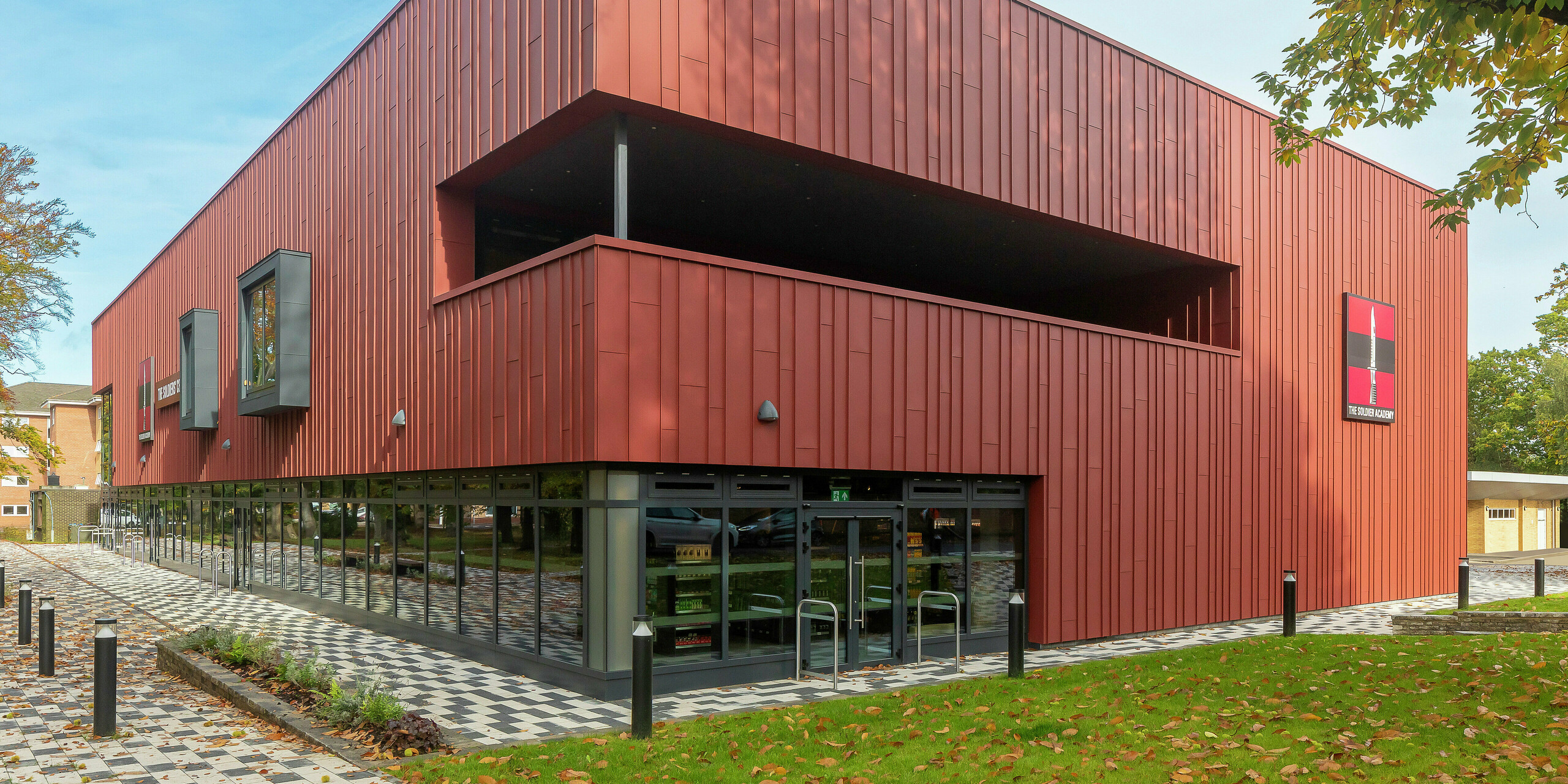 Zeitgemäße Architektur mit PREFALZ: Das Soldiers' Centre in Pirbright überzeugt mit einer modernen Stehfalzfassade aus PREFALZ in P.10 Oxydrot. Die großflächige Verglasung im Erdgeschoss schafft Transparenz und Offenheit, während Fensterrahmen in P.10 Anthrazit stilvolle Akzente setzen. Ein nachhaltiges Baukonzept mit langlebigen PREFA Aluminiumprodukten für höchste Design- und Qualitätsansprüche.