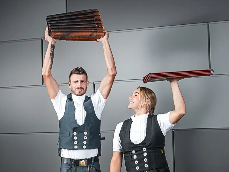 Slika prikazuje dvije osobe koje drže hrpe krovnog materijala. Osoba s lijeve strane drži hrpu krovnih crijepova iznad glave, dok osoba s desne strane drži hrpu aluminijskih krovnih limova, također iznad glave. Obje nose tradicionalnu radnu odjeću, uključujući prsluke i bijele košulje. Pozadina je moderna i minimalistička, sa sivim zidovima. Obje osobe izgledaju usredotočeno i smiješe se.
