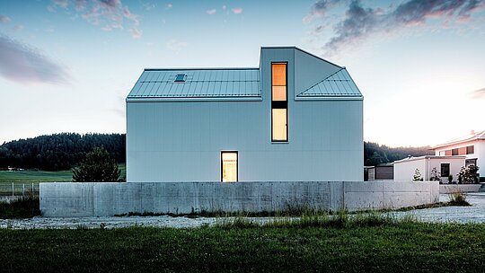Slika prikazuje upečatljivu obiteljsku kuću u Freistadtu, koja se ističe čistim linijama i minimalističkom arhitekturom. Fasada od naboranog aluminija PREFALZ u čistobijeloj boji stvara upečatljivu, geometrijsku strukturu koja zgradi daje moderan i bezvremen izgled. Strmi zabatni krov s ugrađenim krovnim prozorom i visoki prozori osiguravaju obilje dnevnog svjetla te čine unutrašnjost otvorenom i privlačnom. Korištenje PREFALZ-aluminija jamči ne samo estetsku postojanost, već i izvrsnu otpornost na vremenske uvjete i dugovječnost, čime kuća postaje funkcionalna i održiva. Jasna forma i elegantni detalji doprinose sofisticiranom i praktičnom rješenju za modernu stambenu arhitekturu.