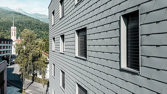 Detaljni prikaz fasade Bernerhausa u Buchsu s precizno postavljenim PREFA zidnim šindrama u P.10 svijetlosivoj boji. Ljuskasta aluminijska površina stvara fino, živopisno uzorkovanje i naglašava jasno urezane prozorske otvore s dubokim otklonima. Lagani, otporni na vremenske uvjete PREFA aluminij osigurava homogenu, oblikovno stabilnu i gotovo bezdržavnu ovojnicu zgrade. Na alpskoj pozadini, visokokvalitetna metalna fasada postaje moderan vizualni naglasak s bezvremenskim dojmom.