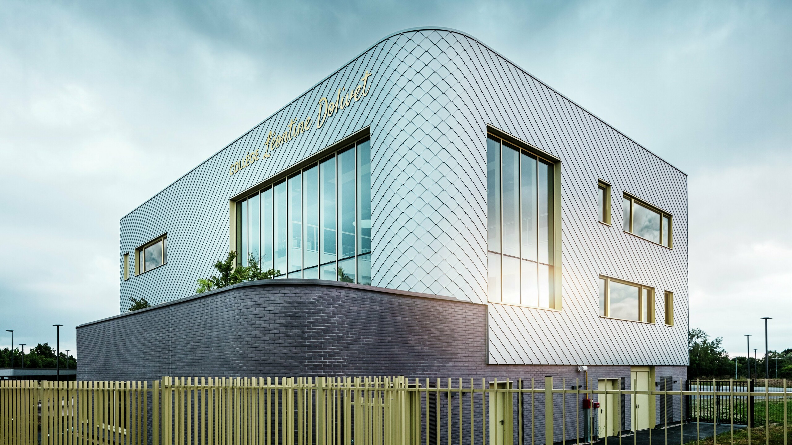 Fotografija prikazuje upečatljiv kut Collège Léontine Dolivet s zakrivljenom aluminijskom fasadom od srebrno-metalnih PREFA zidnih rombova. Velike staklene površine reflektiraju svjetlo, dok žuti okviri prozora stvaraju naglaske. U prvom planu proteže se svijetla ograda uz zgradu, postavljena na tamni klinker u prizemlju. Nebo je oblačno.