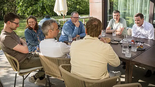 PREFARENZEN 2027 – stručni dijalog u sunčanom vanjskom prostoru vinarske rezidencije: Arhitekti i stručnjaci za dizajn sjede za velikim stolom, duboko uronjeni u raspravu, dok su na stolu raspoređene čaše i mali zalogaji. Scena prikazuje živahnu interakciju i usredotočenu pažnju, okruženu prirodnim zelenilom i sjenama drveća. PREFA aluminijski proizvodi u pozadini predstavljaju dugotrajne i estetski privlačne krovove i fasade. Slika prenosi kako PREFA povezuje dizajn, funkcionalnost i održivost te nadahnjuje kreativne rasprave među arhitektima.