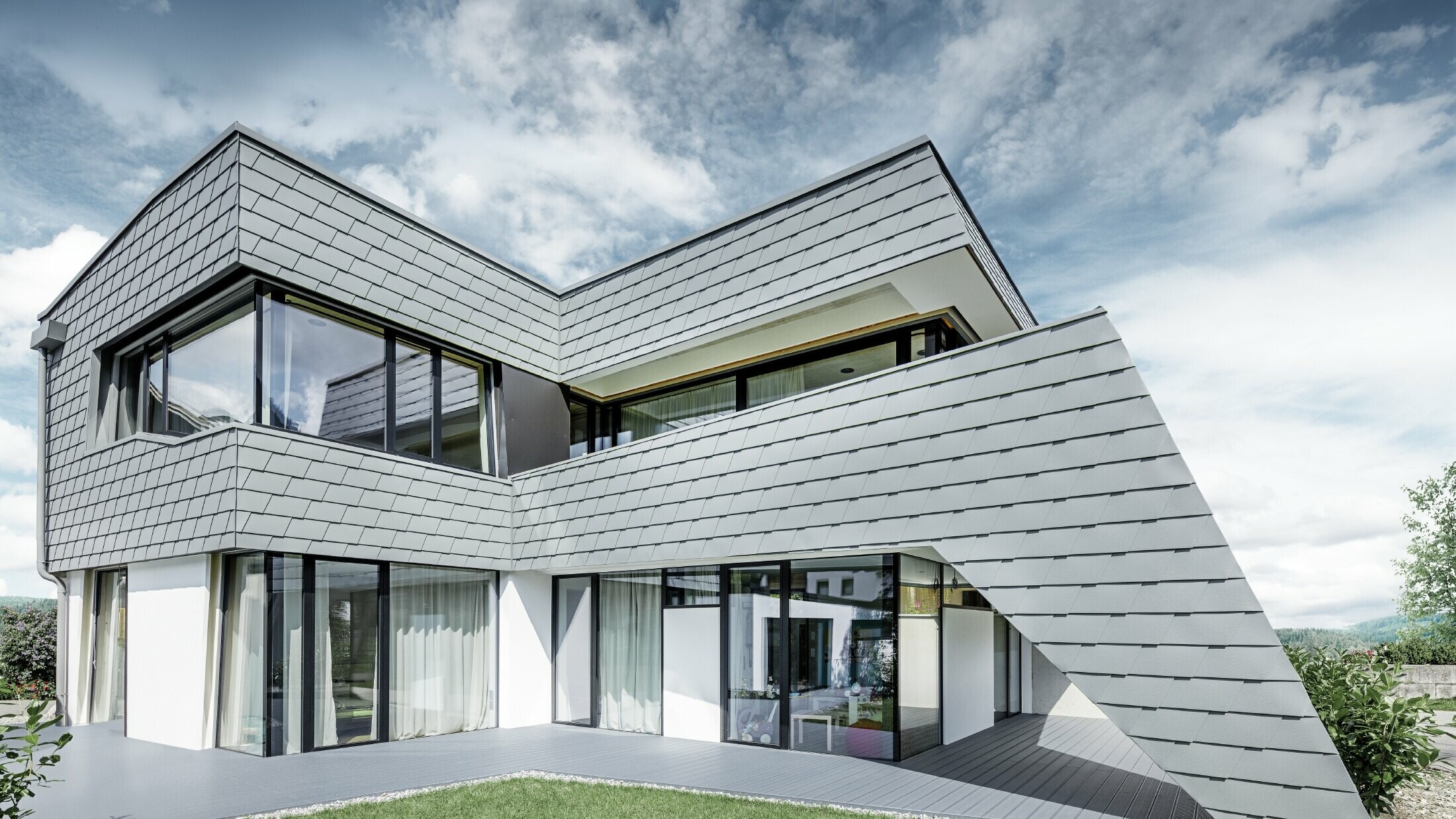 Moderna obiteljska kuća s ravnim krovom, velikim površinama prozora i fasadom iz šindre u svijetlo sivoj boji iz aluminija od PREFA