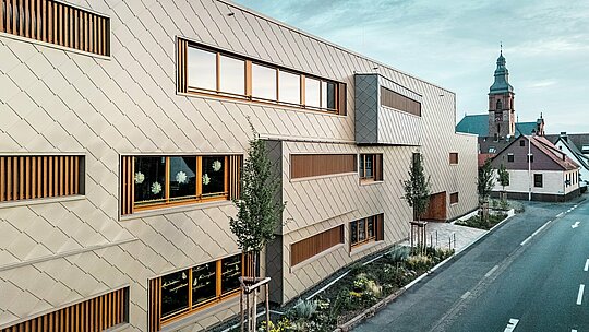 Bočna perspektiva dječjeg vrtića u Alzenauu prikazuje moderno hibridno zdanje u dinamičnom kontrastu s povijesnom gradskom strukturom, dok se u pozadini uzdiže kasnogotička crkva na dvorcu. Fasada od brončano obojenih PREFA zidnih rombova 44 × 44 stvara elegantan, ritmičan uzorak i samouvjereno se ističe u okruženju – bez nametljivosti. Prostrani prozori s vertikalnim drvenim lamelama i strukturirana zelena površina uz zgradu naglašavaju održivi koncept oblikovanja. Kombinacija inovativne aluminijske fasade, ekološke gradnje i urbanističke osjetljivosti čini projekt uzorom odgovorne arhitekture koja spaja tradiciju i budućnost.