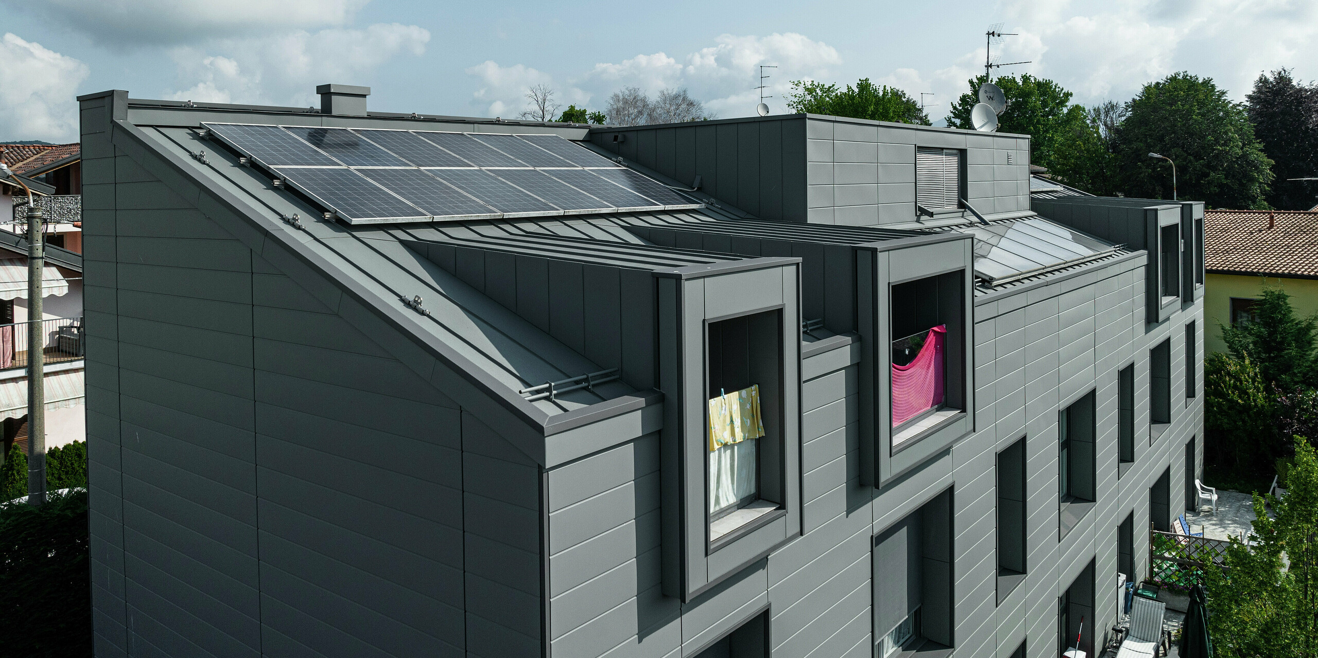 Vogelperspektive auf das Dach einer modernen Wohnanlage in Faloppio, Italien, mit einer robusten Stehfalzverkleidung aus Aluminium und einer nachhaltigen Photovoltaikanlage. Die PREFA Siding-Fassade und das Blechdach bieten nicht nur optische Highlights, sondern auch eine hervorragende Witterungsbeständigkeit und eine nachhaltige Energiequelle durch die PV-Module. Die Kombination aus funktionalen und ästhetischen PREFA Produkten sorgt für eine langlebige und energieeffiziente Lösung.