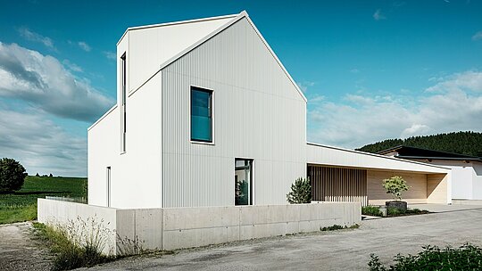 Moderna obiteljska kuća u Freistadtu prikazana iz dinamične perspektive koja ističe čiste linije i upečatljivu arhitekturu. Fasada i krov prekriveni su čistobijelim PREFALZ-aluminijem, stvarajući besprijekornu, modernu i bezvremensku estetiku. Karakteristični elementi dizajna su strmi zabatni krov, velike staklene površine i upečatljivi krovni prozor, dok vertikalni nabori na fasadi zgradi daju zanimljivu teksturu i dubinu. Kontrast s prirodnim okolišem te elegantni drveni elementi na ulazu i garažnim vratima upotpunjuju dizajn. PREFALZ osigurava izuzetno dugotrajnu, lako održivu i otpornu na vremenske uvjete površinu, čineći kuću ne samo estetski privlačnom, nego i funkcionalnom te održivom.