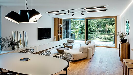 Snimka prikazuje svijetao dnevni i blagovaonski prostor moderne višegeneracijske vile u Budimpešti. Velike staklene stijene otvaraju prostor prema ozelenjenoj terasi, dok svijetli drveni podovi, elegantni dizajnerski namještaj i skladno usklađen koncept rasvjete stvaraju toplu atmosferu. Otvoreni tlocrt povezuje kuhinju, blagovaonicu i dnevni boravak u jedinstvenu cjelinu. Visokokvalitetni interijer nadopunjuje jasnu arhitektonsku formu zgrade, čija je vanjska struktura oblikovana dugotrajnim PREFALZ aluminijskim proizvodima u boji P.10 antracit. Prostor odražava prostranost, modernost i visoku razinu stambenog komfora.