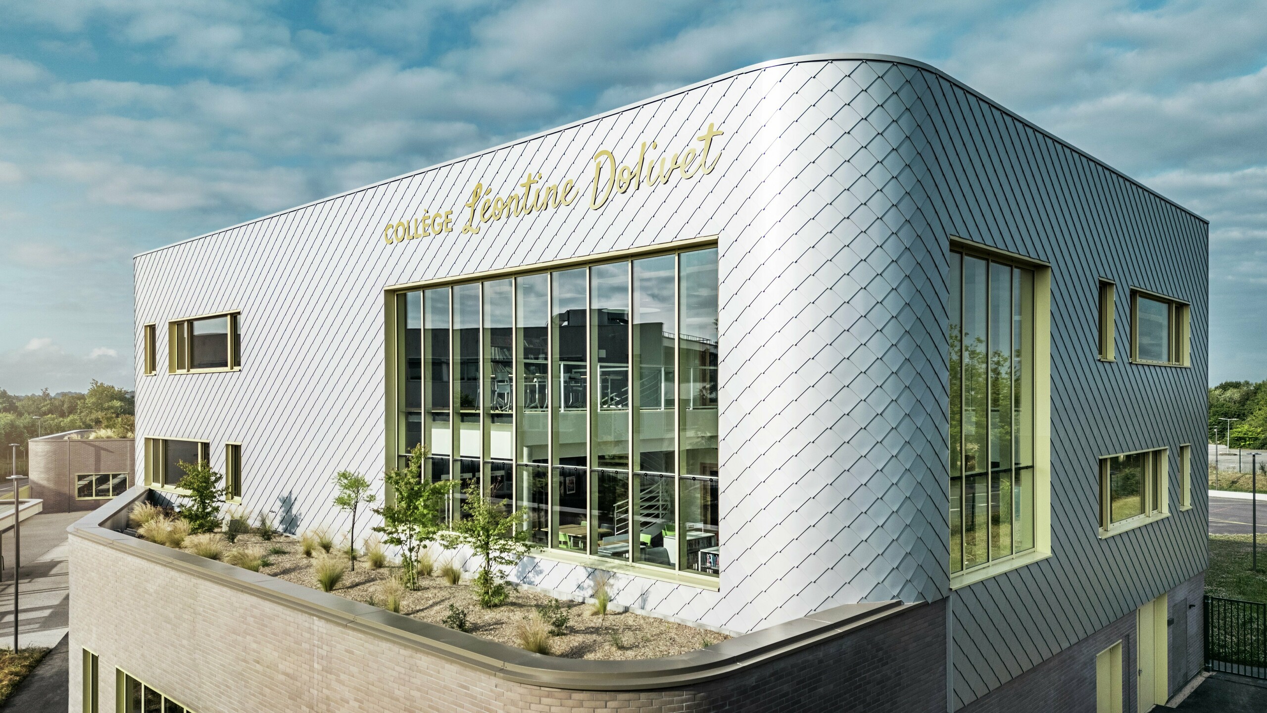 Fotografija prikazuje upečatljiv kut zgrade Collège Léontine Dolivet u Cesson-Sévignéu. Fasada se sastoji od srebrno-metalnih PREFA zidnih rombova, raspoređenih u dijagonalni uzorak koji reflektira svjetlo. Posebno se ističe zakrivljeni oblik gornjeg kata, koji zgradi daje mekani i moderni izgled. Velike staklene površine otvaraju pogled prema unutrašnjosti, dok žuti okviri prozora stvaraju živopisne naglaske. U prvom planu nalazi se zasađeno područje s šljunkom i grmljem, koje arhitekturu skladno nadopunjuje. Lagano oblačno nebo dodatno naglašava metalnu površinu.