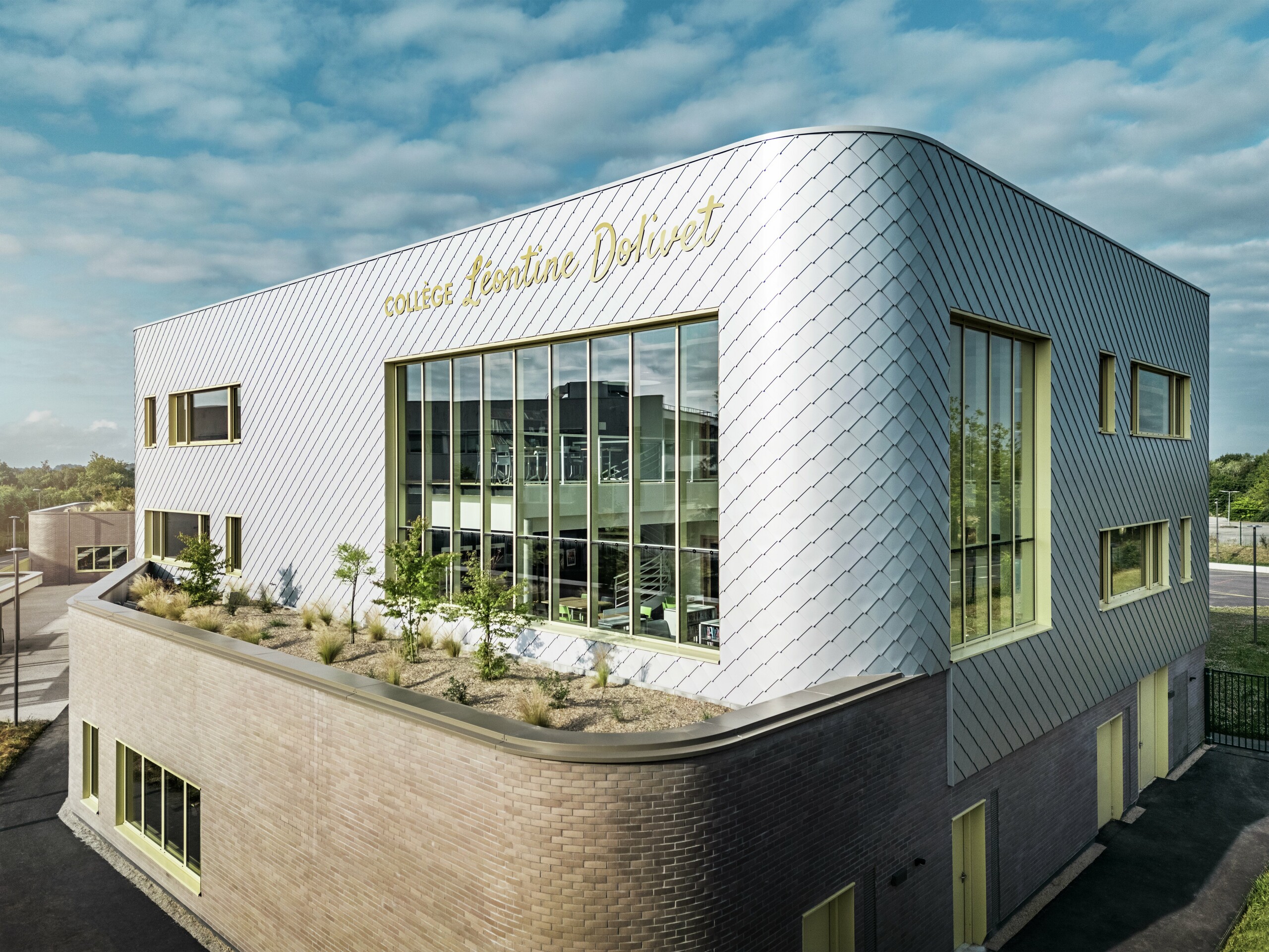 Fotografija prikazuje upečatljiv kut zgrade Collège Léontine Dolivet u Cesson-Sévignéu. Fasada se sastoji od srebrno-metalnih PREFA zidnih rombova, raspoređenih u dijagonalni uzorak koji reflektira svjetlo. Posebno se ističe zakrivljeni oblik gornjeg kata, koji zgradi daje mekani i moderni izgled. Velike staklene površine otvaraju pogled prema unutrašnjosti, dok žuti okviri prozora stvaraju živopisne naglaske. U prvom planu nalazi se zasađeno područje s šljunkom i grmljem, koje arhitekturu skladno nadopunjuje. Lagano oblačno nebo dodatno naglašava metalnu površinu.