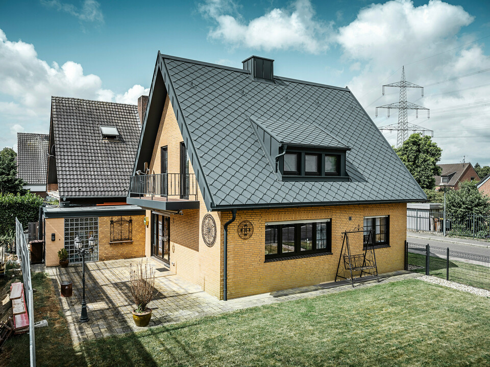 Ein Einfamilienhaus in Tönisvorst mit einem PREFA Dach aus Dachrauten 29×29 in P.10 Anthrazit, das ein herausstechendes Rautenmuster bietet. Schleppgauben, Kamin und der Eingangsbereich sind mit PREFALZ verkleidet, was zusammen mit Ablaufrohr und Hängerinne in P.10 Anthrazit dem Haus einen zeitlosen und eleganten Look verleiht. Das Backsteingebäude mit dem Aluminiumdach harmoniert mit der ruhigen, grünen Umgebung der Siedlung.