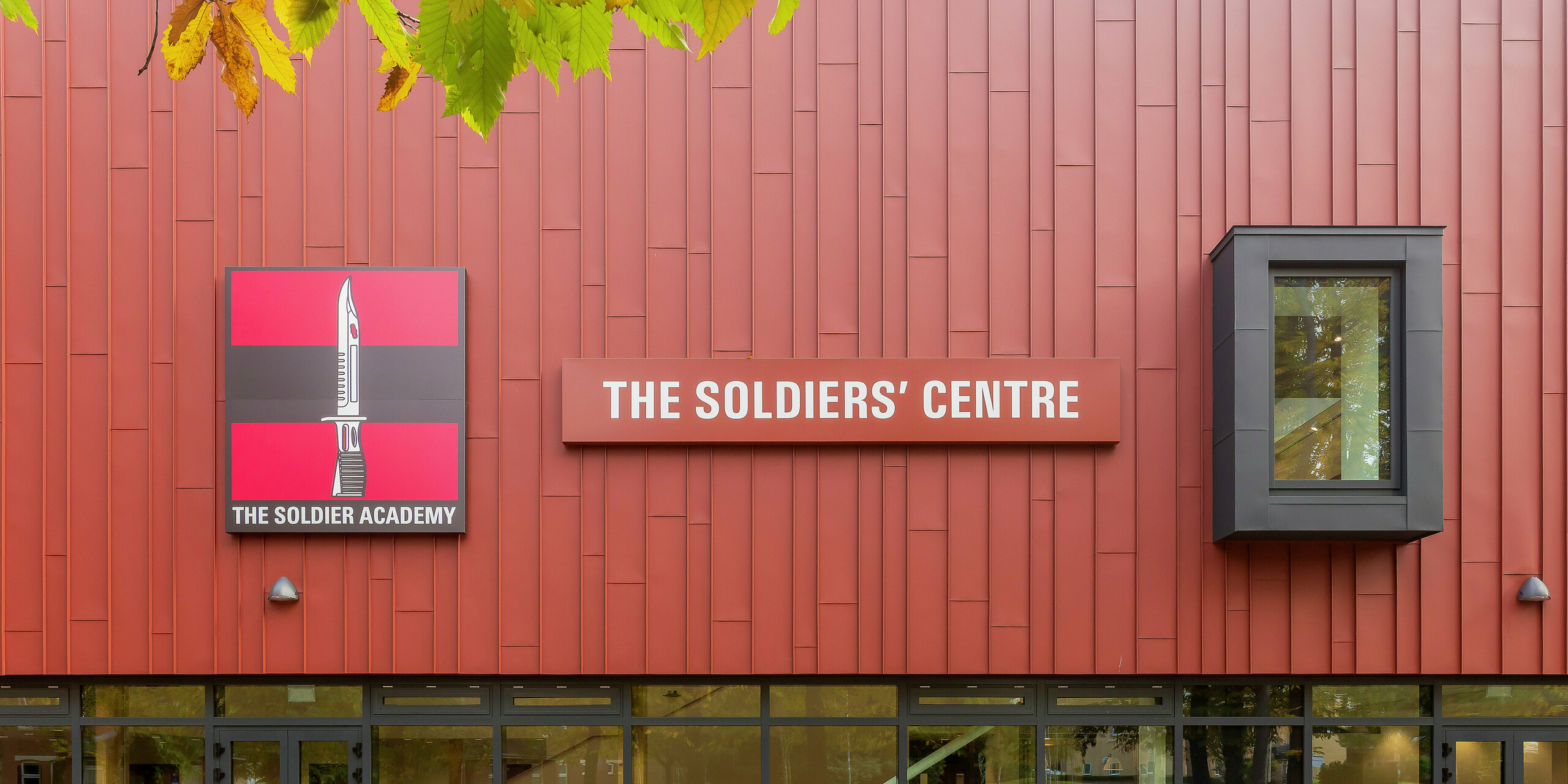 Speziell für die Armee errichtete Sozialeinrichtung mit PREFALZ-Fassade: Die präzise verarbeitete Stehfalzdeckung in P.10 Oxydrot verleiht dem Gebäude ein markantes Erscheinungsbild. Der Eingangsbereich besticht durch großzügige Glasfronten, die für Helligkeit und Transparenz sorgen. Das Logo der 'Soldier Academy' und das auffällige Gebäudeschild setzen klare optische Akzente. Die Fensterrahmen in P.10 Anthrazit runden das Design harmonisch ab. Hochwertige PREFA Aluminiumprodukte für langlebige und ästhetische Architektur.