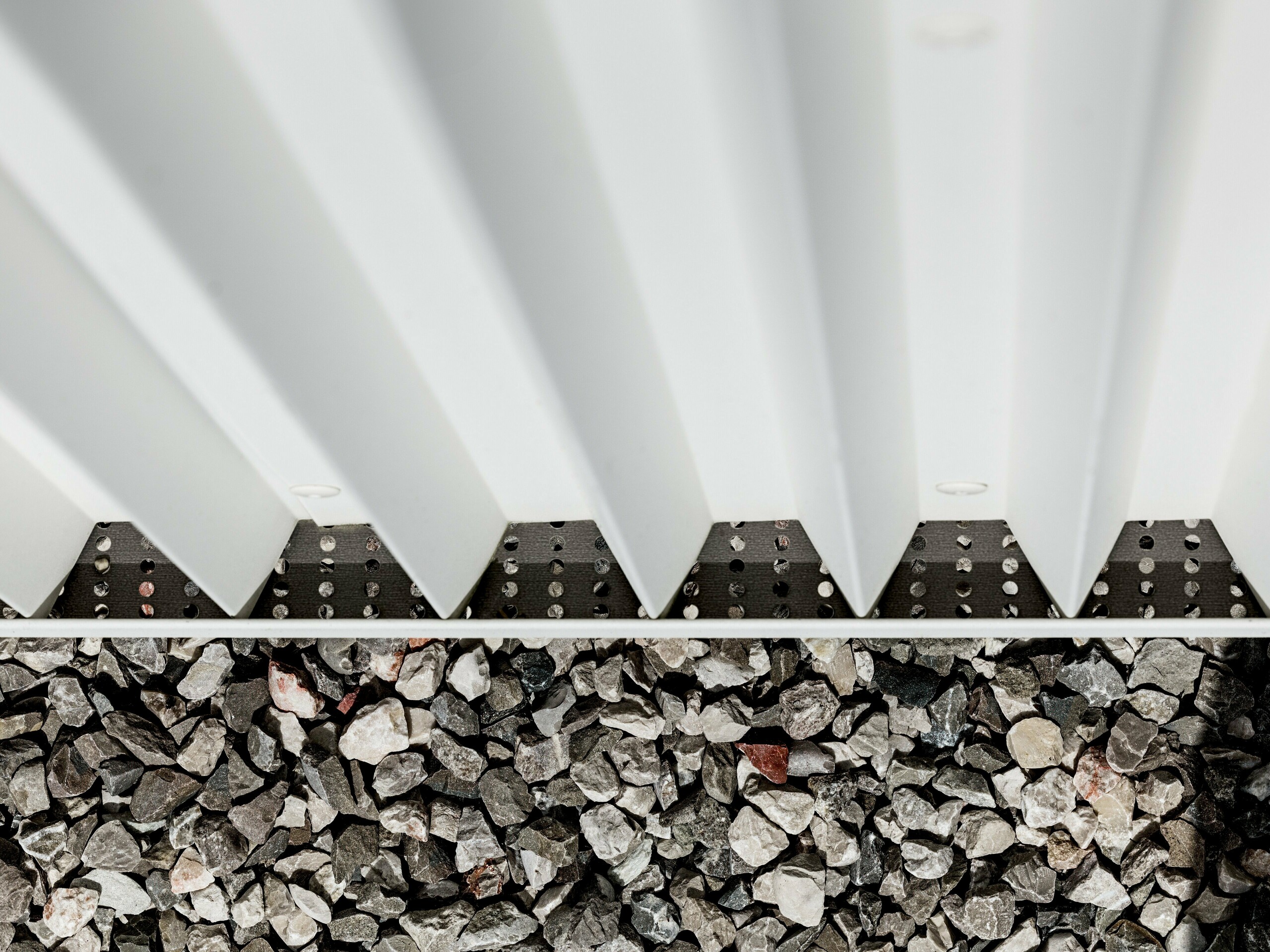 Slika prikazuje detaljan prikaz ventilirane PREFALZ-fasade od čistobijelog aluminija, oblikovane precizno naboranim cik-cak elementima. Ova inovativna tehnika nabiranja ne osigurava samo vizualno privlačnu strukturu, već i izvrsnu ventilaciju te otpornost fasade na vremenske uvjete. Povezivanje elemenata fasade izvedeno je pomoću specijaliziranih, nevidljivih pričvršćivača, što omogućuje posebno čistu i dugotrajnu površinu. Donji dio slike pokazuje šljunčanu podlogu oko zgrade, koja služi za drenažu i stabilizaciju, dodatno naglašavajući funkcionalnost dizajna fasade. Korištenjem PREFALZ-a zadovoljeni su i estetski, i tehnički zahtjevi, što ovaj materijal čini idealnim izborom za modernu i održivu arhitekturu.