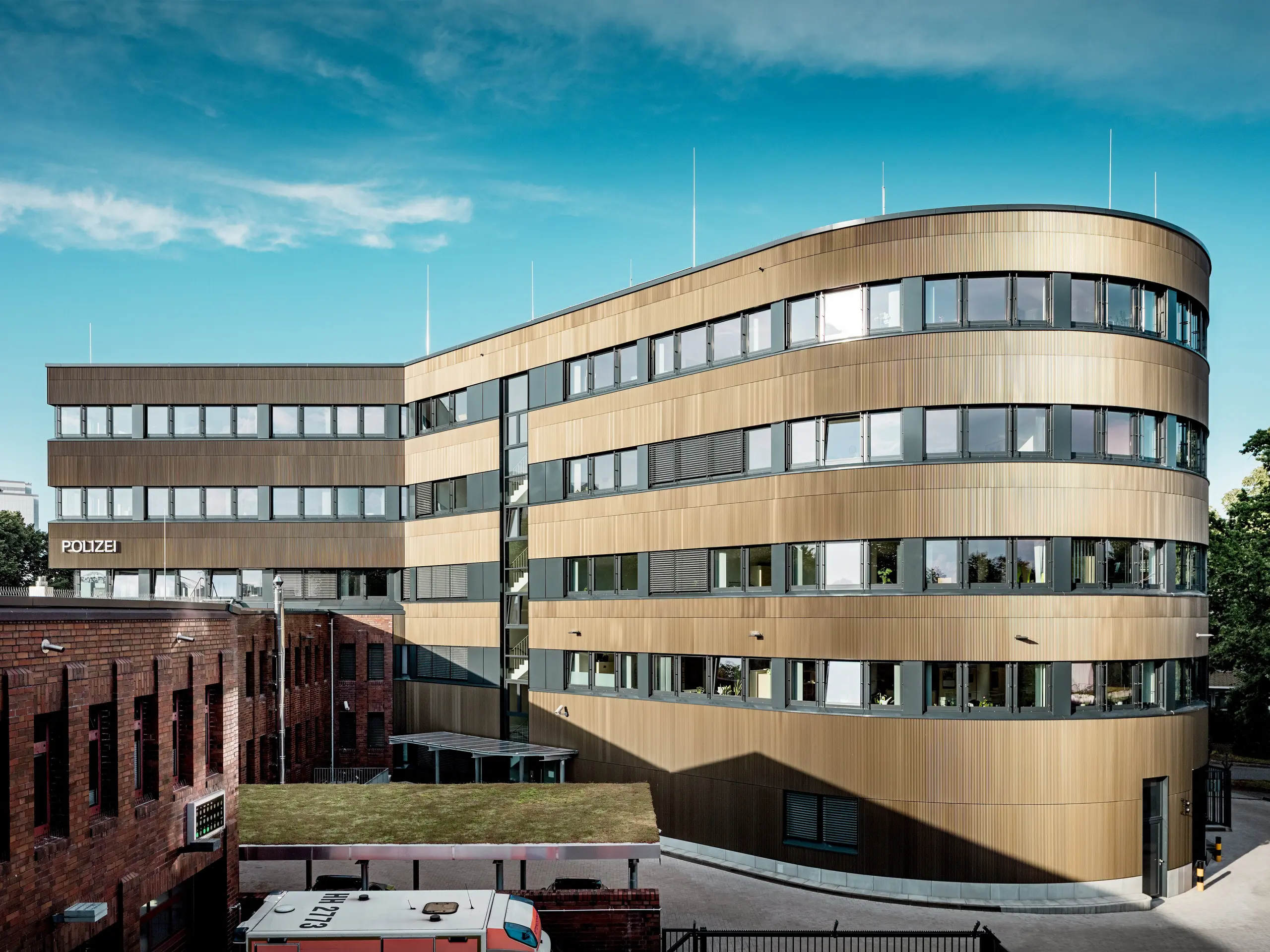 Upečatljiv pogled na cjelokupni objekt Policijske postaje 43 u Hamburgu otkriva zakrivljenu novogradnju koja se svojom PREFA aluminijskom fasadom površine oko 1.100 m² jasno izdvaja od postojeće opečne arhitekture. Fasada je izvedena od ekstrudiranih zupčastih profila u srednje brončanoj nijansi, čija fino strukturirana površina stvara dinamičnu igru svjetla i sjene te objektu daje prepoznatljiv arhitektonski karakter. Zahvaljujući izvrsnoj otpornosti na vremenske utjecaje i koroziju, dugom vijeku trajanja te minimalnom održavanju, PREFA aluminijska rješenja omogućuju održivu, funkcionalnu i estetski zahtjevnu fasadnu arhitekturu.
