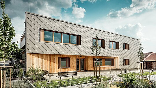 Frontalni prikaz dječjeg vrtića u Alzenauu otkriva pozivajući glavni ulaz s harmonijskom kombinacijom brončano obojenih PREFA zidnih rombova 44 × 44 i vertikalne drvene fasade u prizemlju. Jasno strukturirana aluminijska obloga stvara elegantan uzorak „ljuski“, koji je ritmički prekidan prostranim prozorima s drvenim okvirima i vertikalnim lamelama. U prvom planu, prilagođene staze, cvjetnjaci i klupe naglašavaju visoku kvalitetu boravišnog prostora. Igra dugotrajnog aluminija, prirodnog drva i promišljeno uređene vanjske površine čini zgradu arhitektonskim uzorom održivih, funkcionalnih i estetski vrhunskih obrazovnih objekata s PREFA fasadnim sustavima.