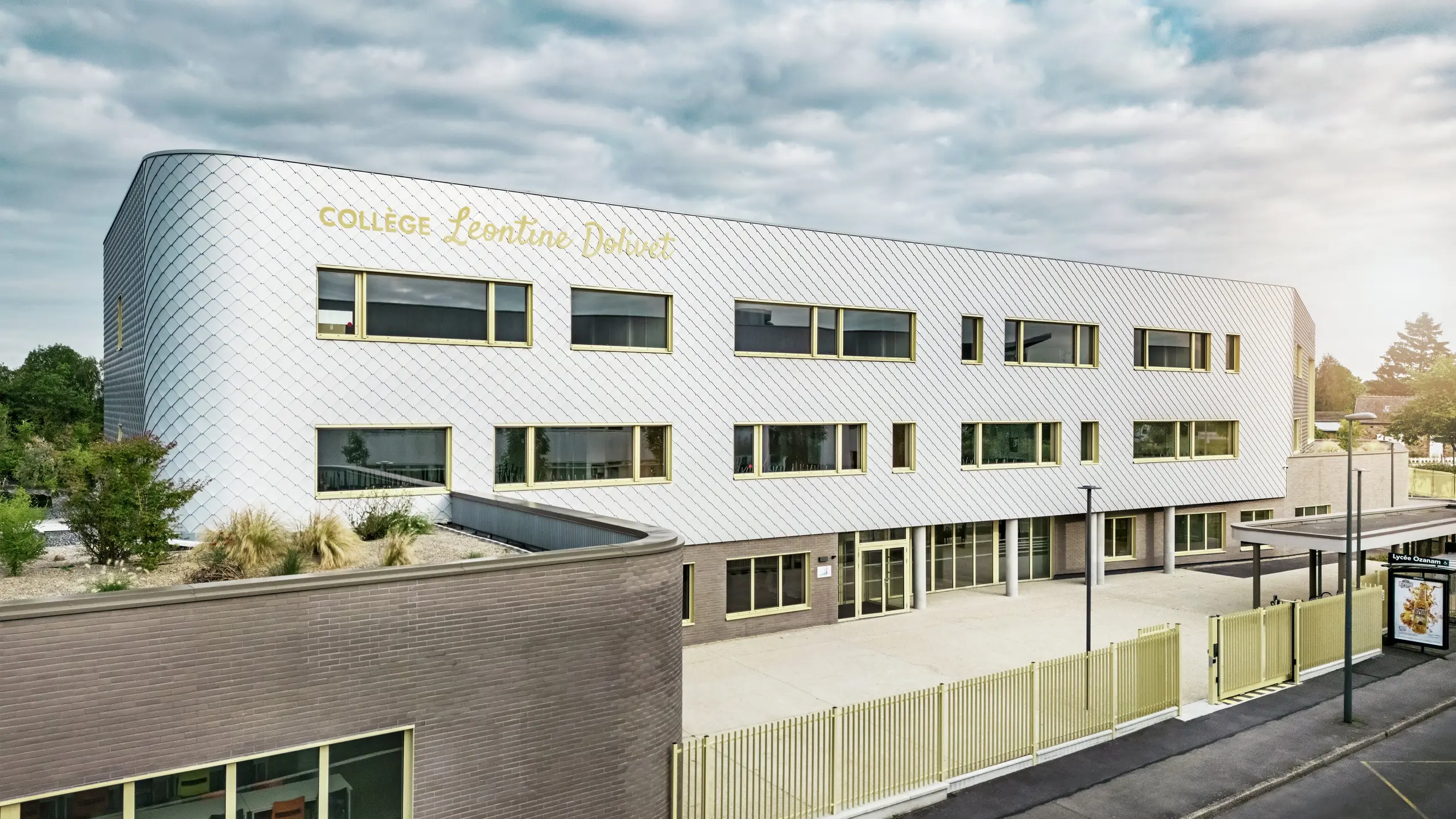 Pogled s prednje strane Collège Léontine Dolivet u Cesson-Sévignéu prikazuje modernu aluminijsku fasadu od PREFA zidnih rombova u srebrno-metalnoj boji. Promjenjiva površina zgradi daje osjećaj lakoće, dok jasne linije i veliki prozori osiguravaju prozirnost. Žuti okviri prozora stvaraju živopisne naglaske. U prvom planu vidljivi su svijetli predprostor, ograda i susjedna ulica. Dugotrajno PREFA rješenje fasade spaja dizajn, otpornost na vremenske uvjete i održivost – arhitektonski privlačan objekt s elegantnim oblikovnim jezikom.