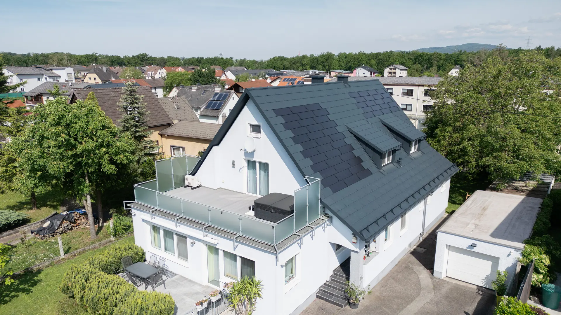 Vogelperspektive auf ein Einfamilienhaus nach der Dachsanierung. Das Dach ist nun mit großen PREFA Solardachplatten und der PREFA Dachplatte R.16 in P.10 Anthrazit gedeckt. Die Solardachplatten sind gleichmäßig verteilt und sorgen für eine moderne, nachhaltige Lösung. Das Gebäude wirkt nun modern und umweltfreundlich. Der Balkon im Vordergrund hat Glasgeländer und befindet sich auf der oberen Etage des Gebäudes.