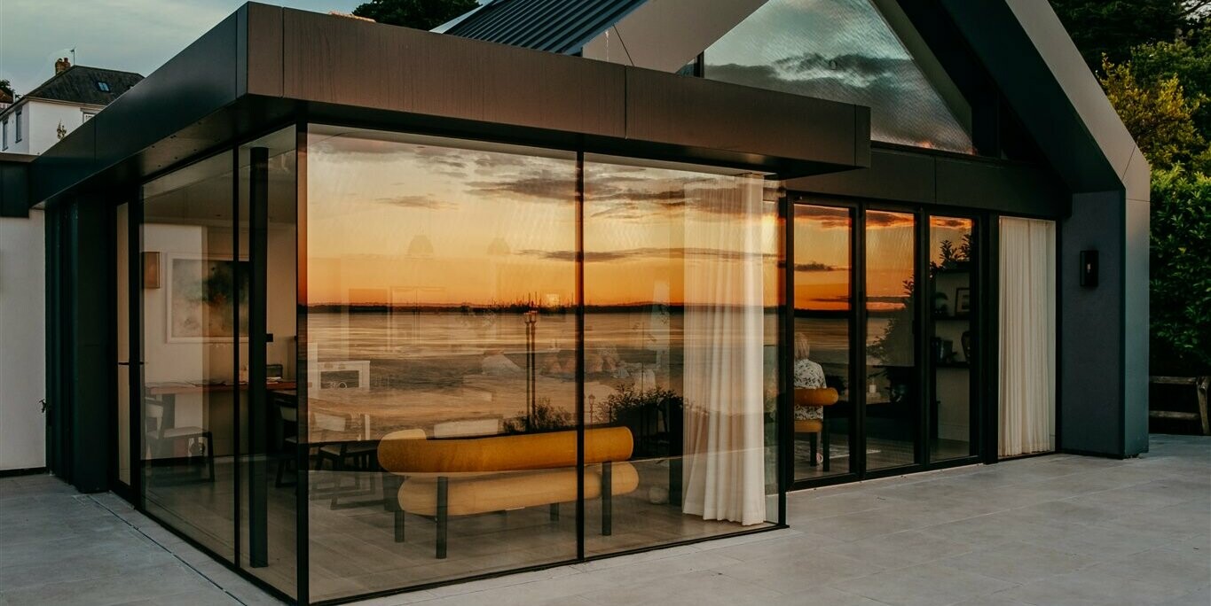 Nahaufnahme von der Terrasse eines luxuriösen Bungalows mit Meerblick auf der Isle of Wight. In den großzügigen Fenstern spiegelt sich der traumhafte Ausblick des Sonnenuntergangs an der Südküste Englands wider. Die großen Glasflächen sind umrahmt von PREFALZ in P.10 Dunkelgrau. Durch die Fenster sind Teile der modernen Innenarchitektur der Luxusimmobilie zu erkennen.