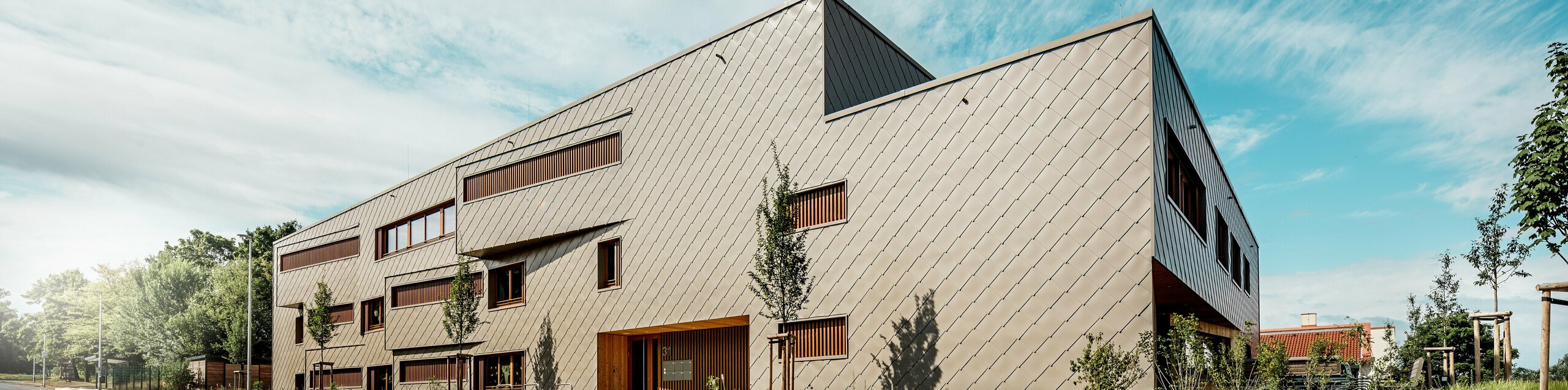 Iz upečatljive perspektive odozdo dječji vrtić u Alzenauu prikazuje se sa svojom izraženom, dinamičnom linijskom arhitekturom i visokokvalitetnom PREFA aluminijskom fasadom u brončanoj boji. Dijagonalni raspored zidnih rombova 44 × 44 daje građevini jasnu strukturu i elegantnu, homogenu površinu. Pomaknuti prozori i vertikalne drvene lamele stvaraju ritmičke akcenta, dok reducirani oblikovni jezik odaje dojam moderne suzdržanosti. Kombinacija dugotrajnog aluminija, prirodnog drva i promišljenog zelenog pojasa uz ulicu naglašava visoke standarde održivosti i građevinske kvalitete. Ova slika dojmljivo pokazuje kako funkcionalna arhitektura u kombinaciji s PREFA proizvodima može estetski impresionirati i postaviti urbane standarde.