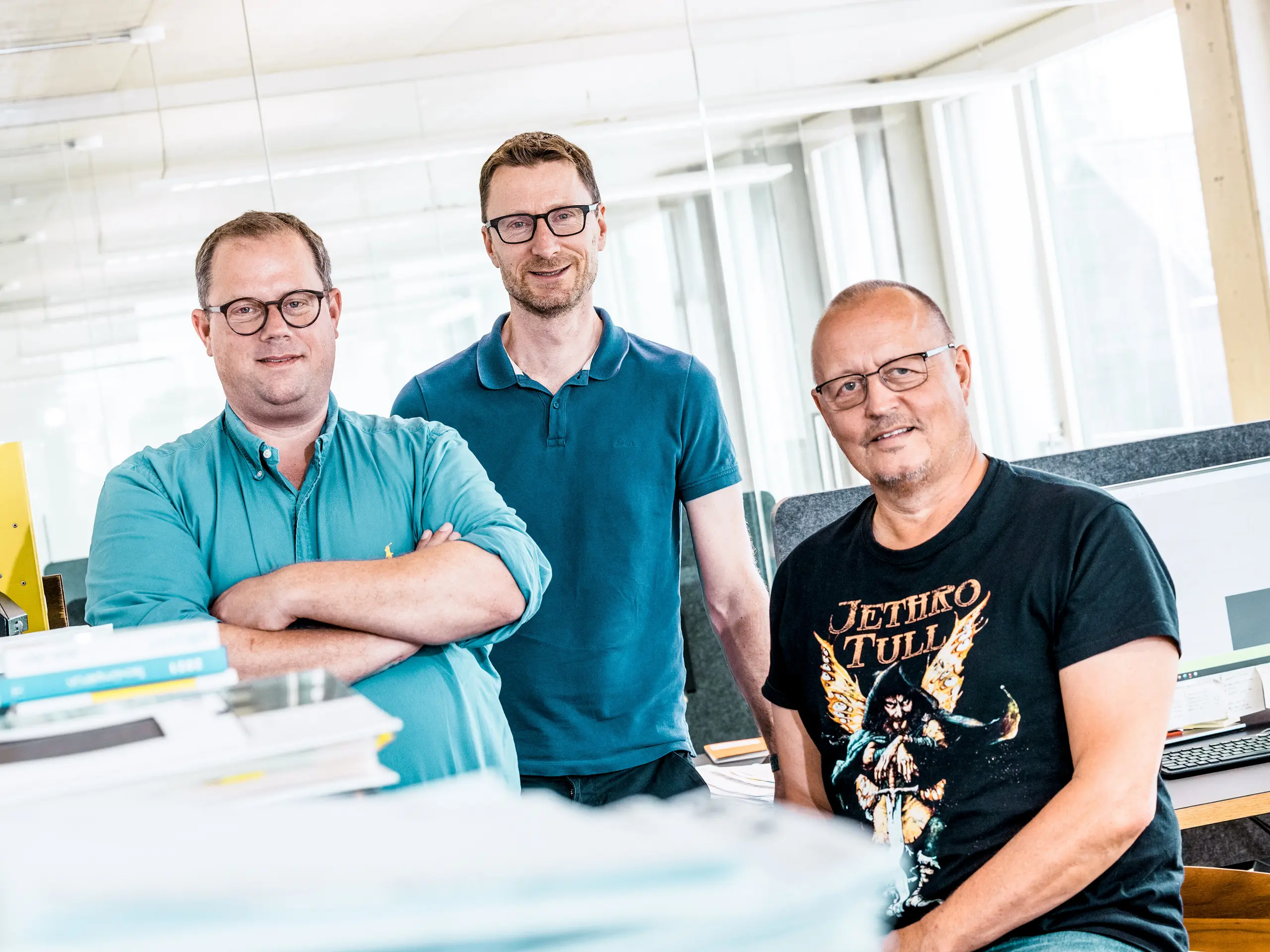 Timska fotografija arhitekata iz studija Kaundbe u njihovom modernom uredu: tri arhitekta stoje samouvjereno ispred svojih radnih mjesta i tehničkih dokumenata. U prvom planu nalaze se hrpe planova i projektne mape, a u pozadini se vide ekrani računala s konstrukcijskim crtežima. Scena prenosi dojam stručnosti, suradnje i tehničke preciznosti – temeljnih vrijednosti u planiranju i realizaciji fasadnih rješenja s PREFA aluminijskim proizvodima.