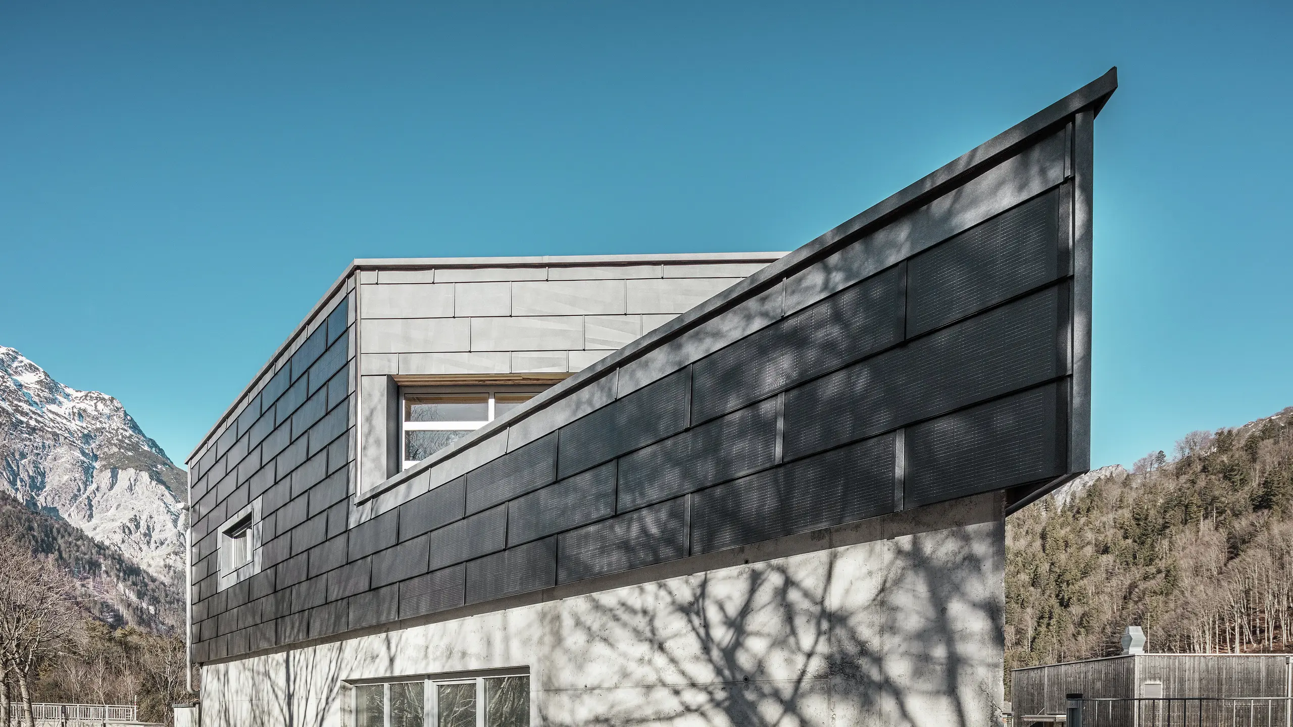 Poslovna zgrada limarske radionice RÜMA u Vorarlbergu impresionira svojom modernom fasadom od PREFA solarnih krovnih ploča i vidljivog betona. Okružena planinama i drvećem čiji se sjena iscrtava na zidu, stoji pod čistim plavim nebom. Oštri kut zgrade i PREFA fasadni paneli FX.12 dodatno naglašavaju upečatljiv izgled.