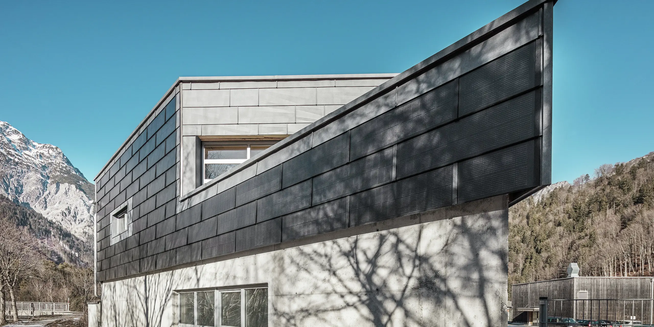 Poslovna zgrada limarske radionice RÜMA u Vorarlbergu impresionira svojom modernom fasadom od PREFA solarnih krovnih ploča i vidljivog betona. Okružena planinama i drvećem čiji se sjena iscrtava na zidu, stoji pod čistim plavim nebom. Oštri kut zgrade i PREFA fasadni paneli FX.12 dodatno naglašavaju upečatljiv izgled.