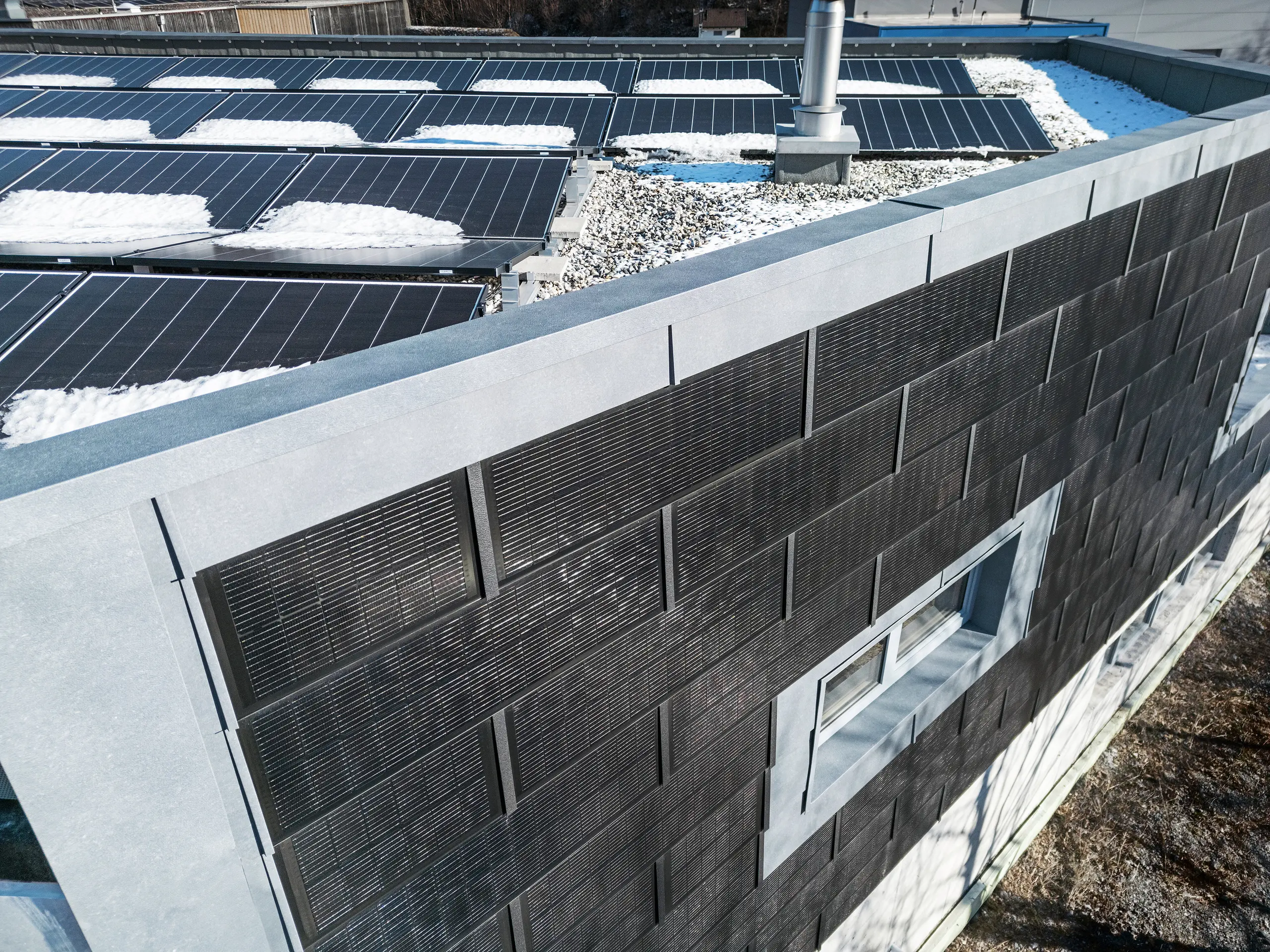 Pogled iz zraka prikazuje fasadu limarske radionice RÜMA u Vorarlbergu s sivim fasadnim panelima FX.12 te inovativnim PREFA solarnim krovnim pločama s integriranim fotonaponskim modulima. U prvom planu vidi se dijelom snijegom prekriven ravan krov s dodatnim fotonaponskim modulima.