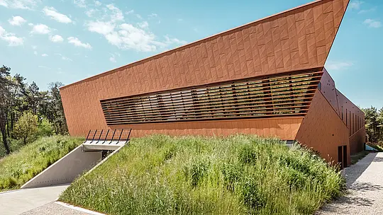 Upečatljiva crpna stanica za pitku vodu WML u Lottumu ima izražajno nagnut vrh zgrade i dinamično oblikovanu PREFA Siding.X aluminijsku fasadu u toplom izgledu hrđe. Fasetirana površina stvara živahnu igru svjetla i sjene, dok horizontalne lamelne trake stvaraju arhitektonsku dubinu. Uklopljena u prirodno oblikovanu padinu, zgrada djeluje poput skulpturalne orijentacijske točke u prostoru. Dugotrajni, lagani i reciklabilni PREFA aluminijski paneli odlikuju se visokom otpornošću na vremenske uvjete, stabilnošću oblika te održivim fasadnim omotačem koji zahtijeva minimalno održavanje.