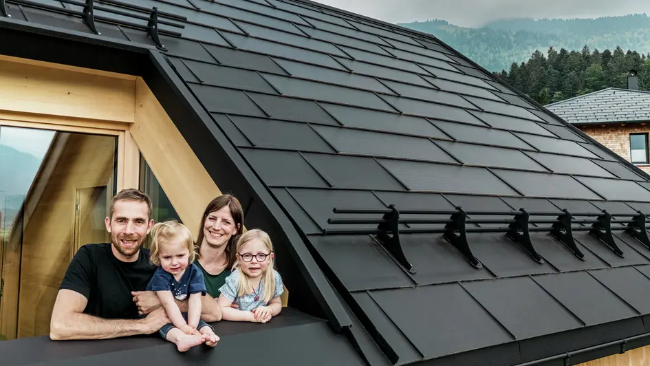 Dvojica vlasnika kuće i njihova djeca uživaju u pogledu s loggie svoje novogradnje, koja je opremljena elegantnim PREFA solarnim krovnim pločama u boji P.10 Black.