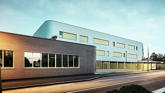 Abendansicht des Collège Léontine Dolivet in Cesson-Sévigné. Im Vordergrund verläuft eine Straße mit klarer Markierung, daneben ein flacher Gebäudeteil mit großen Glasfenstern. Dahinter erhebt sich der Hauptbau mit geschwungener Aluminiumfassade aus PREFA Wandrauten in Silbermetallic, die im sanften Licht changiert. Mehrere rechteckige Fenster und ein heller Zaun strukturieren die Ansicht. Die Szene ist von Straßenlaternen beleuchtet, der Himmel zeigt ein kühles Blau – ein moderner, eleganter Blickfang.