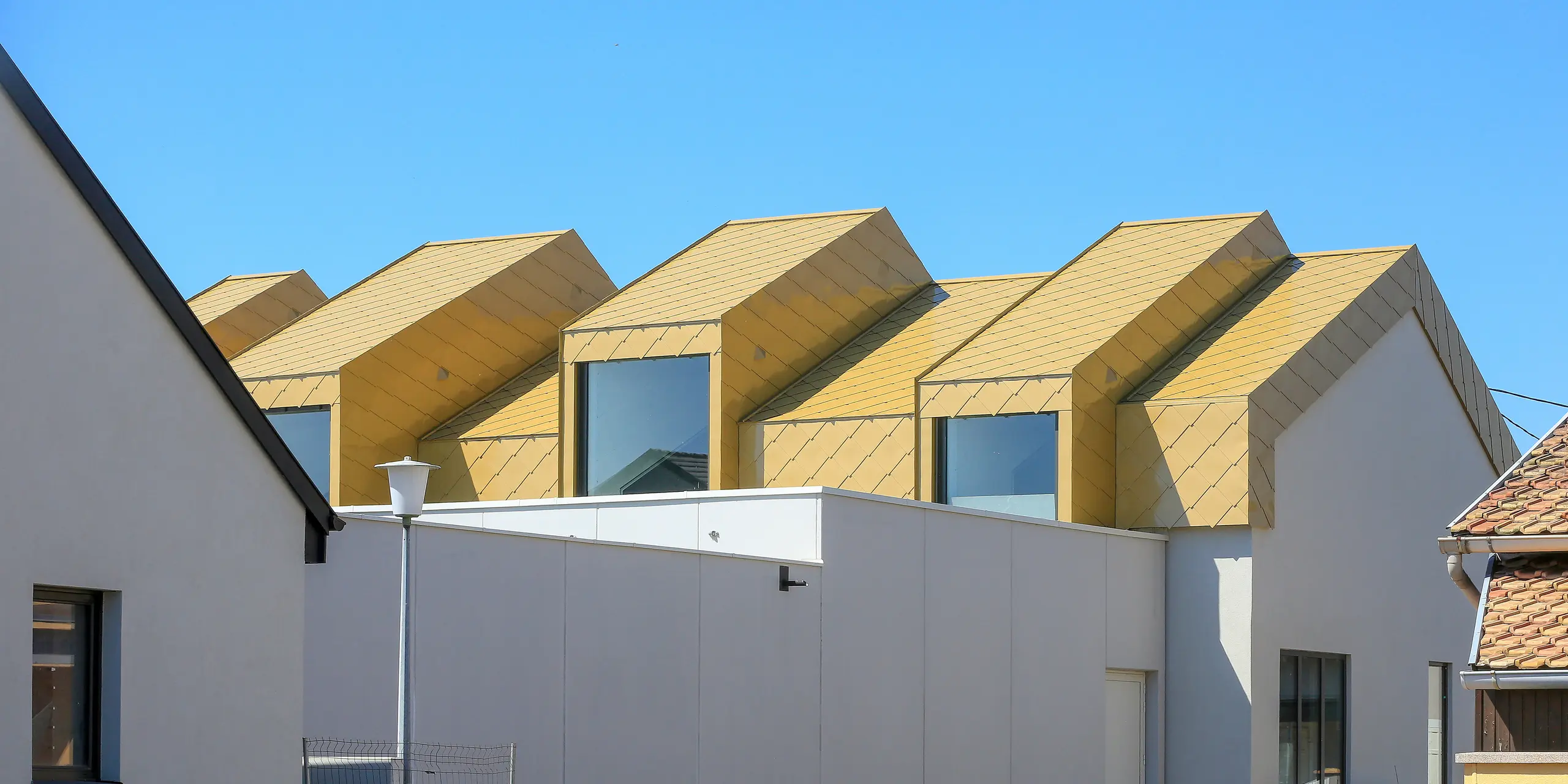 Blick aus der Distanz auf das Gemeinde- und Kulturzentrum Fessenheim (FRA). Die Perspektive auf das Gebäude mit den glänzenden PREFA Dach- und Wandrauten 44 × 44 in Sparkling Gold ist von der Straße.