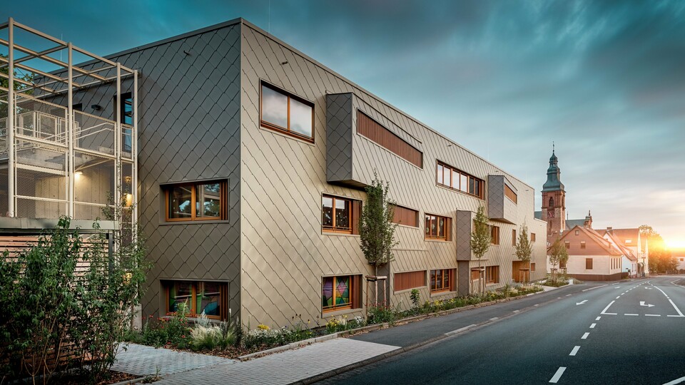 U mekom svjetlu zalaska sunca dječji vrtić u Alzenauu pokazuje se u svom najdojmljivijem izdanju. Fasada od brončano obojenih PREFA zidnih rombova 44 × 44 reflektira večernje svjetlo i naglašava visokokvalitetnu, strukturiranu površinu modernog hibridnog objekta. Jasno raščlanjena arhitektura s pomaknutim trakama prozora i vertikalnim drvenim lamelama stvara uzbudljiv kontrast povijesnoj crkvi u pozadini. S lijeve strane slike filigranska metalna konstrukcija vanjskog stubišta osigurava laganu transparentnost. Uređeni zeleni pojas uz zgradu dodatno naglašava ekološku orijentaciju. Ova slika na dojmljiv način povezuje suvremenu arhitektonsku kulturu, održivu PREFA aluminijsku tehnologiju i skladno uklapanje u povijesnu gradsku sliku Alzenaua.