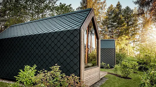 Minimalistička tiny kuća Secret Koi Gardena na jezeru Natterer See smještena je u zelenilu i uokvirena toplim večernjim svjetlom. Krov i fasada u potpunosti su obloženi sitnim PREFA krovnim i zidnim rombovima od aluminija u P.10 antracitnoj boji, čija precizna struktura nalik ribljoj koži stvara izražajne naglaske.  Trajni i vremenski otporni PREFA aluminij pouzdano štiti arhitekturu te se savršeno usklađuje s drvom, staklom i okolnom prirodom.