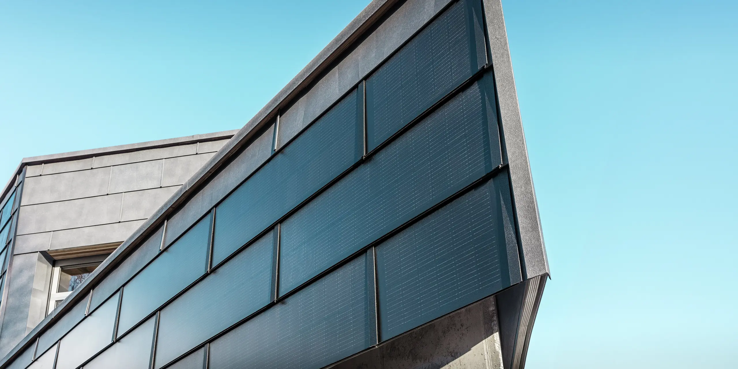 Moderna poslovna fasada limarske radionice RÜMA u Vorarlbergu s PREFA solarnim krovnim pločama od aluminija i integriranim fotonaponskim modulima, u kombinaciji s PREFA fasadnim panelima FX.12, za održivu proizvodnju energije i dinamičan dizajn fasade. Snimka prikazuje kut zgrade izbliza. U pozadini se vidi vedro plavo nebo.