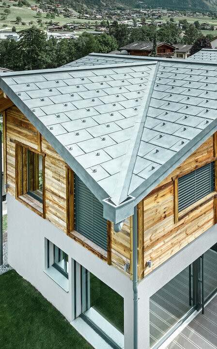 Švicarska kuća s aluminijskim krovom tvrtke PREFA. Postavljena je krovna ploča R.16 u kameno sivoj. Na gornjoj etaži postavljena je rustikalna drvena fasada.