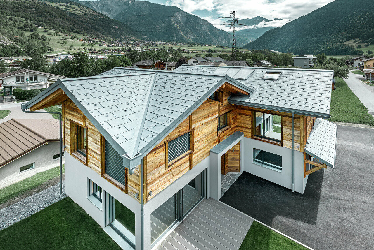 Švicarska kuća s aluminijskim krovom tvrtke PREFA. Postavljena je krovna ploča R.16 u kameno sivoj. Na gornjoj etaži postavljena je rustikalna drvena fasada.