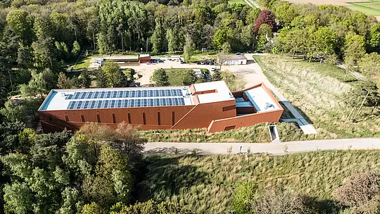 Zračna snimka crpne stanice za pitku vodu WML u Lottumu kao upečatljive infrastrukturne građevine, skladno uklopljene u zeleni krajolik šuma i livada. Izduženi volumen zgrade s izražajnom PREFA Siding.X aluminijskom fasadom u toplom izgledu hrđe djeluje poput skulpturalne orijentacijske točke u prostoru. Vizualni naglasci su jasno strukturirani oblik zgrade, integrirani fotonaponski moduli na krovu te fino fasetirana površina fasade. Dugotrajni, lagani i reciklabilni PREFA aluminijski paneli odlikuju se visokom otpornošću na vremenske uvjete, održivom učinkovitošću i fasadnim omotačem koji zahtijeva minimalno održavanje.