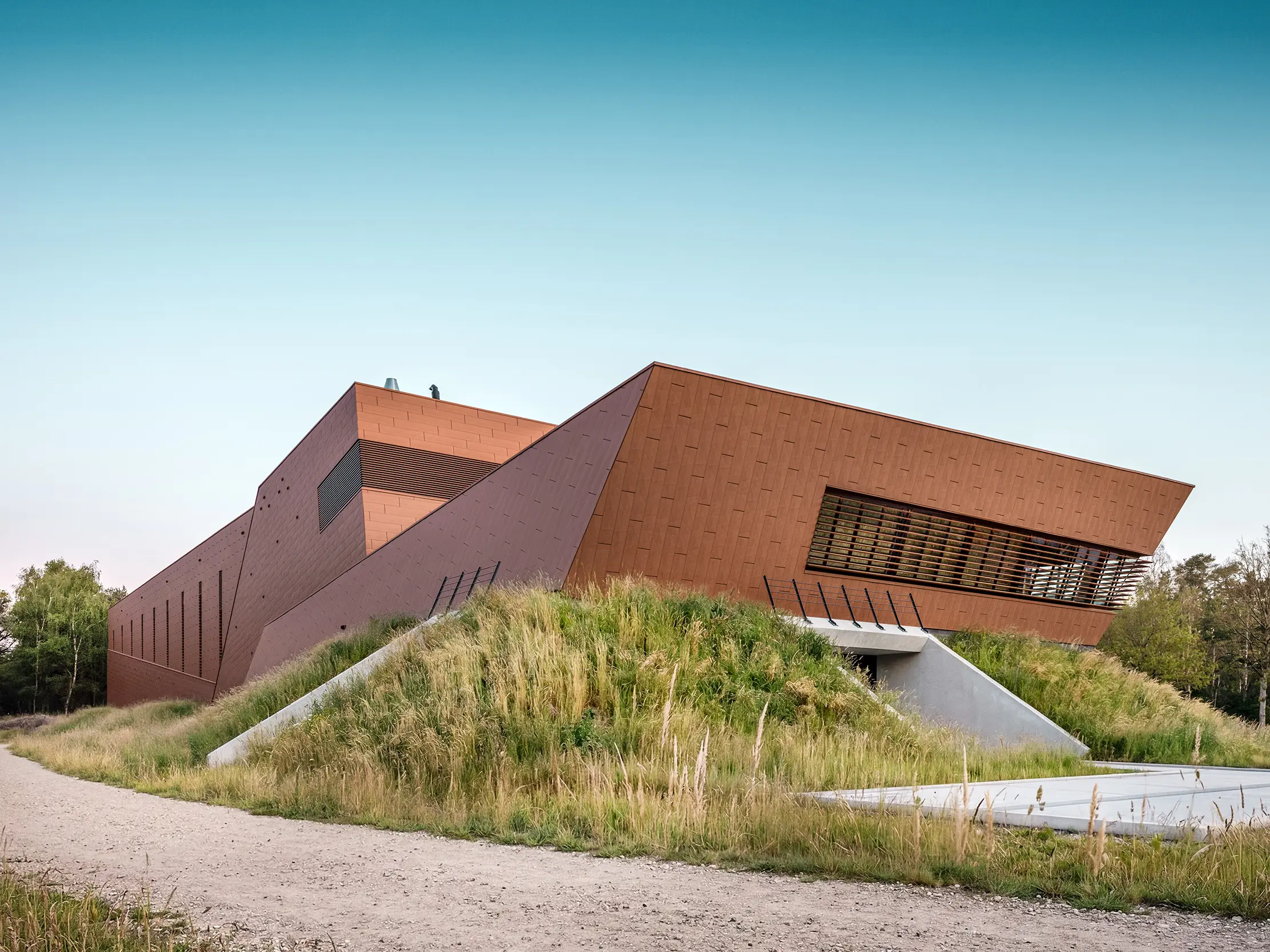 Arhitektonski izražajna crpna stanica za pitku vodu WML u Lottumu sa skulpturalno oblikovanim, presavijenim volumenima zgrade, uklopljena u prirodni krajolik padine. Upečatljiva PREFA Siding.X aluminijska fasada u toplom izgledu hrđe privlači poglede svojom fasetiranom, živopisnom površinom te dojmljivo poigrava svjetlom i sjenom. Horizontalne lamele i precizne linije naglašavaju dinamičnu geometriju. Lagani, dugotrajni i potpuno reciklabilni PREFA aluminijski paneli predstavljaju vrhunsku otpornost na vremenske uvjete, održivu gradnju i fasadni omotač koji zahtijeva minimalno održavanje.