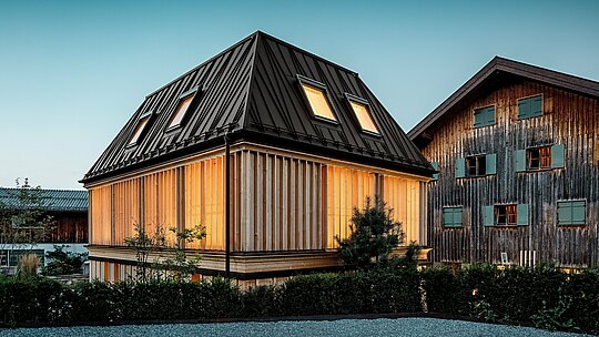 Nagrađivani „Badehaus“ hotela Hirschen u Schwarzenbergu u atmosferičnom večernjem svjetlu. U prvom planu dominira moderni krov s četiri vode s upečatljivim PREFA falcom u boji P.10 orah smeđa – pravo arhitektonsko istaknuto djelo koje plijeni pažnju svojom nekonvencionalnom linijom i nagibom od 60 stupnjeva. Fasada od vertikalnih, etažom visokih drvenih lamela skladno nadopunjuje povijesni ansambl i spaja moderan minimalizam s graditeljskom tradicijom Bregenzerwalda. U pozadini je vidljivo zaštićeno glavno zdanje – snažan kontrast starog i novog. Izvrsno primjer budućnosti orijentirane gradnje s visokokvalitetnim PREFA materijalima.