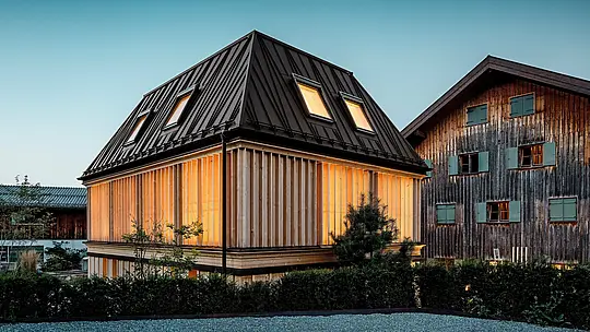 Nagrađivani „Badehaus“ hotela Hirschen u Schwarzenbergu u atmosferičnom večernjem svjetlu. U prvom planu dominira moderni krov s četiri vode s upečatljivim PREFA falcom u boji P.10 orah smeđa – pravo arhitektonsko istaknuto djelo koje plijeni pažnju svojom nekonvencionalnom linijom i nagibom od 60 stupnjeva. Fasada od vertikalnih, etažom visokih drvenih lamela skladno nadopunjuje povijesni ansambl i spaja moderan minimalizam s graditeljskom tradicijom Bregenzerwalda. U pozadini je vidljivo zaštićeno glavno zdanje – snažan kontrast starog i novog. Izvrsno primjer budućnosti orijentirane gradnje s visokokvalitetnim PREFA materijalima.