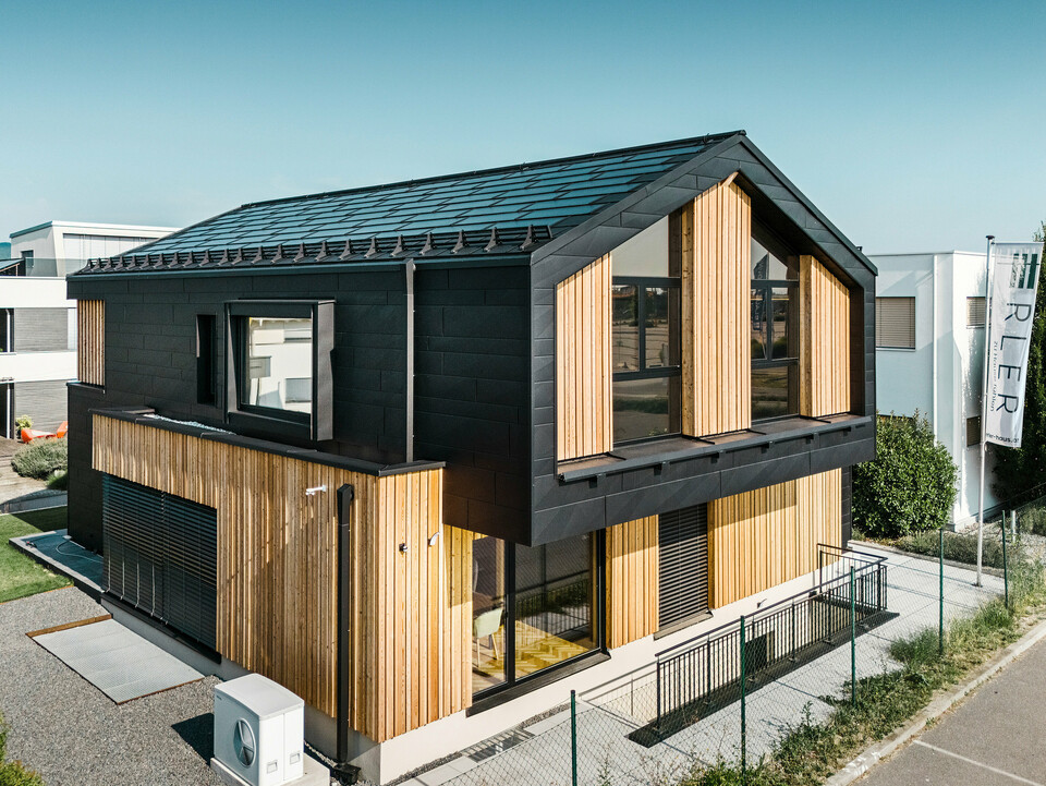 Sprednja stran vzorčnega doma Autark v centru za montažne hiše Blaue Lagune, Vösendorf (AT). Streha je pokrita s črnimi solarne strešne plošče PREFA in strešne plošče R.16 v visokokakovostni P.10-premazu. Fasada je sestavljena iz kombinacije navpičnih lesenih elementov in PREFA Siding.X v P.10 črna barvi, kar stavbi daje sodoben in trajnosten videz. Na desnem robu slike plapola zastava podjetja Erler Bau GmbH.