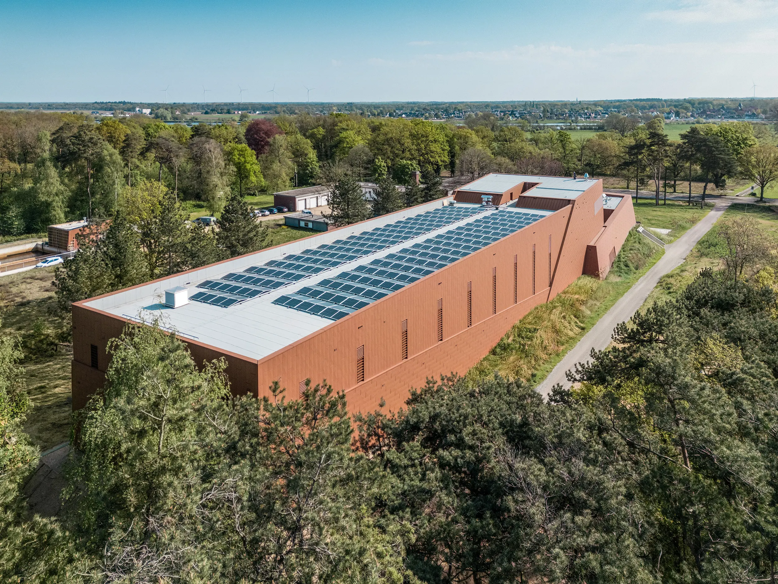 Zračna snimka crpne stanice za pitku vodu WML u Lottumu, uklopljene u prostrani krajolik drveća i polja. Izdužena struktura zgrade impresionira upečatljivom PREFA Siding.X aluminijskom fasadom u toplom izgledu hrđe i krovom ispunjenim fotonaponskim modulima kao održivim vizualnim elementom. Fino strukturirani omotač zgrade živahno reflektira svjetlost i naglašava moderni oblikovni izraz. Lagani, dugotrajni i reciklabilni PREFA aluminijski paneli osiguravaju visoku otpornost na vremenske uvjete, energetsku učinkovitost i arhitekturu usmjerenu prema budućnosti koja zahtijeva minimalno održavanje.