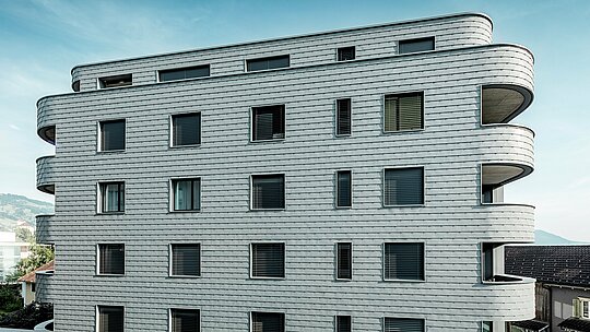 Frontalni pogled na Bernerhaus u Buchsu s nježno zaobljenim uglovima zgrade i kontinuiranom fasadom od PREFA zidnih šindra u P.10 svijetlosivoj boji. Aluminijska fasada položena poput ljuski stvara živopisni uzorak i naglašava horizontalni ritam zakrivljenih balkonskih traka. Precizno ugrađena prozorska otvora skladno se uklapaju u homogenu površinu. Zahvaljujući laganom, otpornom na vremenske uvjete i dugotrajnom PREFA aluminiju, nastaje gotovo bezdržavna, oblikovno stabilna ovojnica zgrade s upečatljivom prisutnošću na alpskoj pozadini.