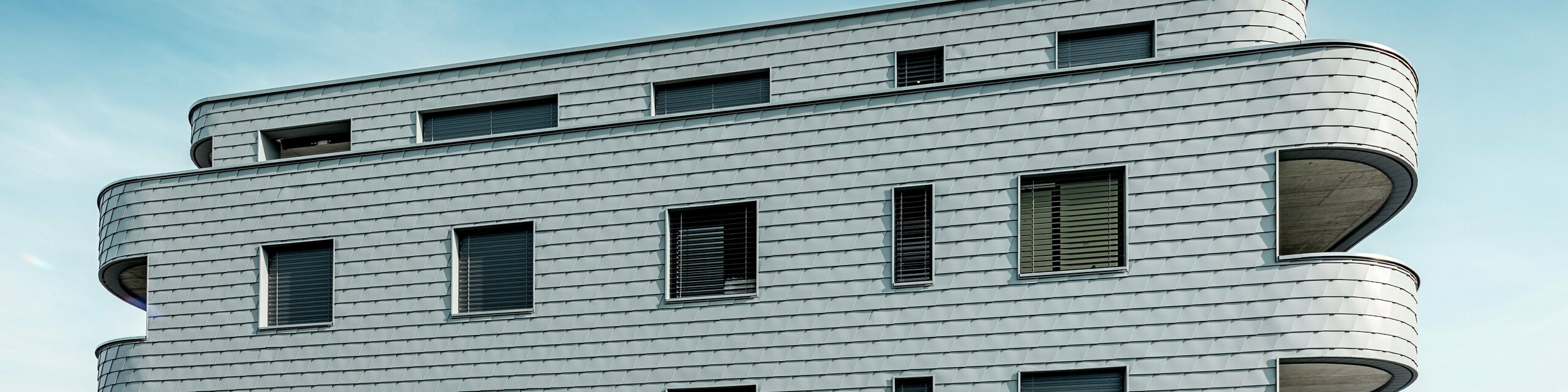 Frontalni pogled na Bernerhaus u Buchsu s nježno zaobljenim uglovima zgrade i kontinuiranom fasadom od PREFA zidnih šindra u P.10 svijetlosivoj boji. Aluminijska fasada položena poput ljuski stvara živopisni uzorak i naglašava horizontalni ritam zakrivljenih balkonskih traka. Precizno ugrađena prozorska otvora skladno se uklapaju u homogenu površinu. Zahvaljujući laganom, otpornom na vremenske uvjete i dugotrajnom PREFA aluminiju, nastaje gotovo bezdržavna, oblikovno stabilna ovojnica zgrade s upečatljivom prisutnošću na alpskoj pozadini.