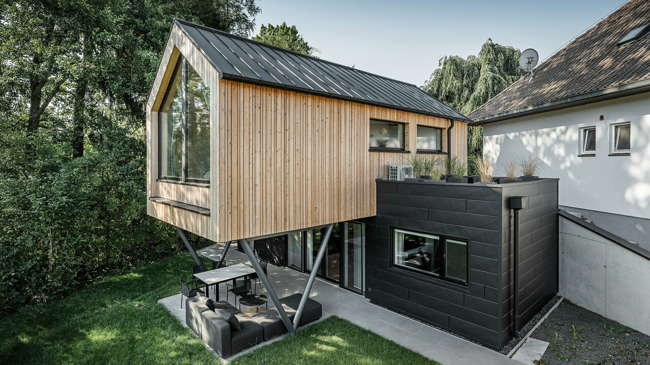 Das 'Woodstock Chalet 5' beeindruckt mit einer eindrucksvollen Kombination aus natürlichen Holzoberflächen und den modernen PREFA Siding.X Fassadenelementen in Anthrazit. Die überhängende Holzstruktur des Obergeschosses ragt markant über die dunklen Fassadenelemente, die das Erdgeschoss verkleiden, und erzeugt so ein Spiel aus Licht und Schatten. Die klaren Linien und die robuste Konstruktion der Siding.X Elemente ergänzen das innovative Design des Gebäudes und bieten einen spannenden visuellen Kontrast zur umgebenden Vegetation und dem traditionellen Gebäude im Hintergrund. Am Dach des Chalets wurde das PREFALZ Dachsystem in Form einer gleichmäßigen Stehfalzdeckung verlegt. Am rechten Rand des Erdgeschosses ist das PREFA Dachentwässerungssystem zu erkennen, das analog zur Aluminiumfassade in Anthrazit gehalten ist. Für die Dachentwässerung wurden der Wasserfangkasten gefolgt vom Ablaufrohr montiert.