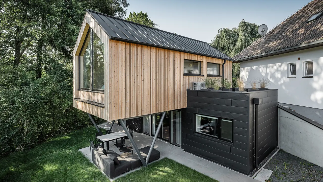 Das 'Woodstock Chalet 5' beeindruckt mit einer eindrucksvollen Kombination aus natürlichen Holzoberflächen und den modernen PREFA Siding.X Fassadenelementen in Anthrazit. Die überhängende Holzstruktur des Obergeschosses ragt markant über die dunklen Fassadenelemente, die das Erdgeschoss verkleiden, und erzeugt so ein Spiel aus Licht und Schatten. Die klaren Linien und die robuste Konstruktion der Siding.X Elemente ergänzen das innovative Design des Gebäudes und bieten einen spannenden visuellen Kontrast zur umgebenden Vegetation und dem traditionellen Gebäude im Hintergrund. Am Dach des Chalets wurde das PREFALZ Dachsystem in Form einer gleichmäßigen Stehfalzdeckung verlegt. Am rechten Rand des Erdgeschosses ist das PREFA Dachentwässerungssystem zu erkennen, das analog zur Aluminiumfassade in Anthrazit gehalten ist. Für die Dachentwässerung wurden der Wasserfangkasten gefolgt vom Ablaufrohr montiert.