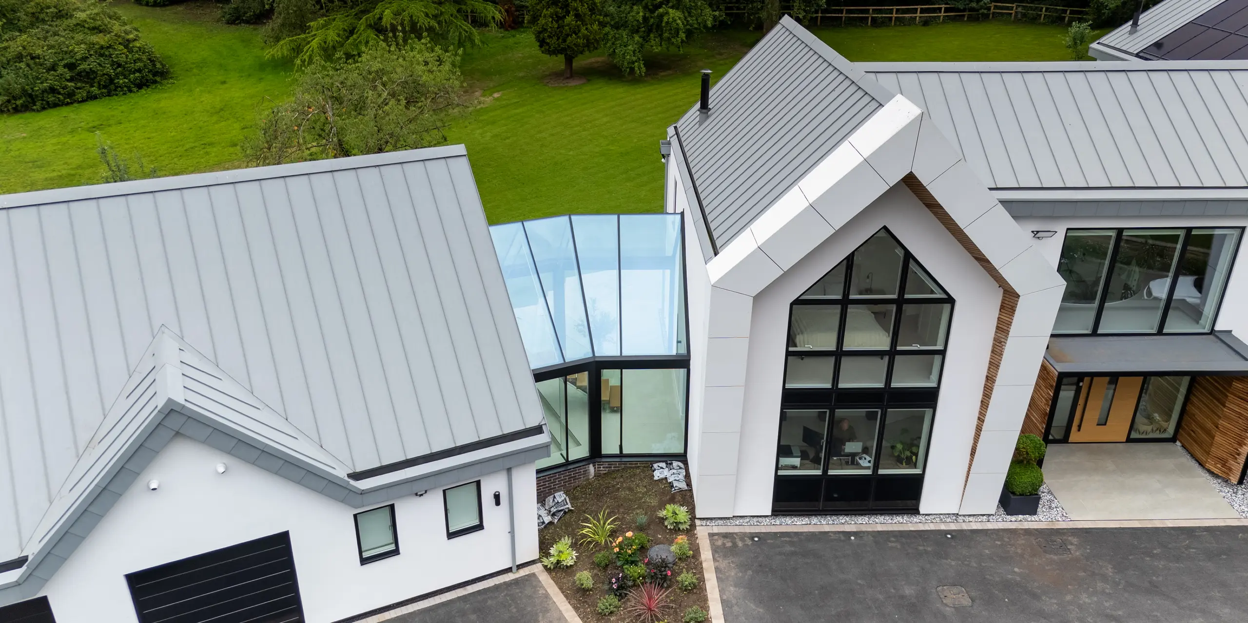 Die Luftaufnahme zeigt eine beeindruckende Ansicht der modernen Villa in Draycott In The Clay, mit ihrem markanten PREFALZ-Dach in Patinagrau. Die symmetrische Stehfalzstruktur des Dachs hebt das Gebäude hervor und fügt sich nahtlos in die Umgebung ein. Besonders auffällig ist die Glasverbindung zwischen den beiden Gebäudeteilen, die den Raum mit Licht durchflutet und das innovative Design betont. Diese Verbindung zeigt eine harmonische Mischung aus modernem Bau und umweltbewusster Architektur, mit Solarpanels, die das Dach ergänzen und die nachhaltige Ausrichtung des Hauses unterstreichen.